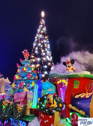 Mickey y Pluto en una carroza con forma de tren que echa humo frente al árbol de Navidad de Disneyland Paris iluminado