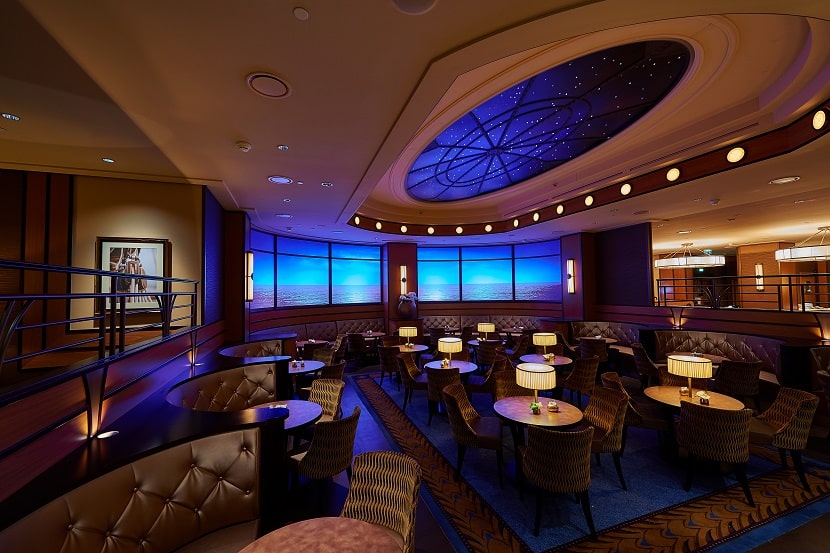 Local decorado con pequeñas mesas redondas, sillas, sofás y alfombras azules, con una iluminación tenue creando un ambiente acogedor en el Compass Lounge del hotel Newport Bay Club de Disneyland Paris