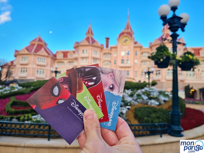 Entradas a Disneyland Paris en papel Mano sujetando cuatro entradas de colores frente al Disneyland Hotel