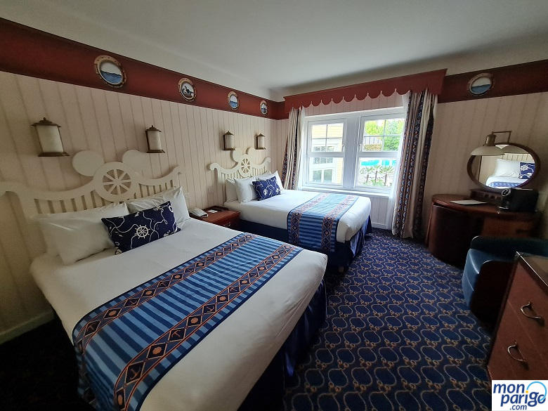 Habitación del hotel Newport Bay Club de Disneyland Paris con dos camas de matrimonio, suelo de moqueta, mesa de escritorio, un espejo, un sillón, una cafetera, etc.