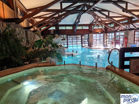 Jacuzzi en un espacio climatizado junto a una piscina interior con un tobogán y grandes cristaleras con luz natural