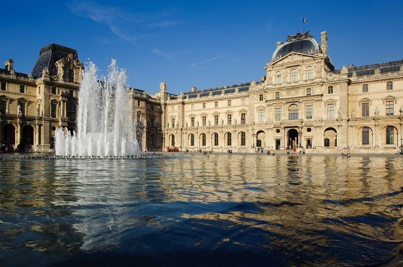Exterior del Museo del Louvre con fuentes de agua en primer plano y reflejo en el estanque de la Cour Napoléon.